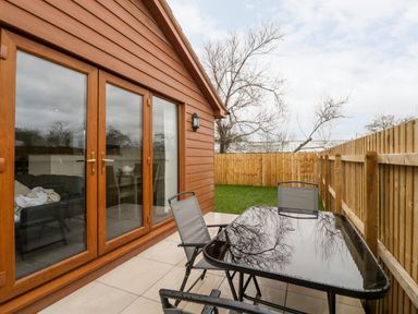 A fenced patio area with a black table and three chairs next to a wooden house with glass doors at Bluebell Lodge Meadow view lodges Brean