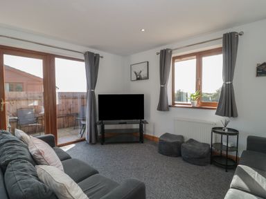 A living room with a sofa television two poufs a side table and large windows with curtains at Bluebell Lodge Meadow view lodges in Brean