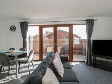 A living room with a gray sofa and cushions a glass dining table with chairs a wall clock and a tv on a stand with glass doors leading to a fenced patio at Bluebell Lodge Meadow view lodges in Brean