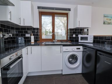 A kitchen with white cabinets black countertops black tiled backsplash a washing machine window and appliances at Bluebell Lodge Meadow view lodges in Brean
