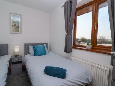 A bedroom with two single beds a nightstand with a lamp and a window with grey curtains at Bluebell Lodge Meadow view lodges in Brean