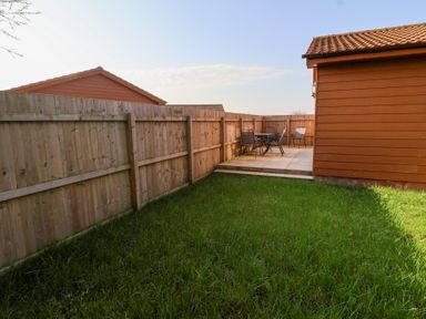 A small fenced backyard with grass and a patio area with table and chairs at Bluebell Lodge Meadow view lodges in Brean