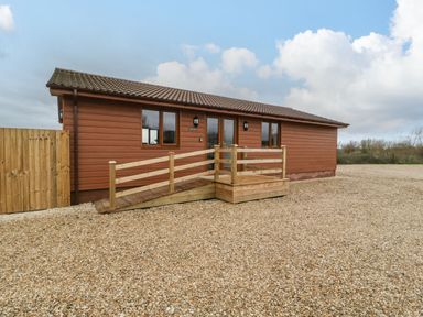 An exterior view of a single story wooden building with a sloped roof and a wooden ramp surrounded by gravel at Delphine Lodge Meadow View Lodges in Brean
