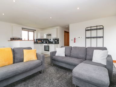 A living room with gray sofas and yellow and white cushions next to a kitchen with white cabinets and black backsplash at Delphine Lodge Meadow View Lodges in Brean
