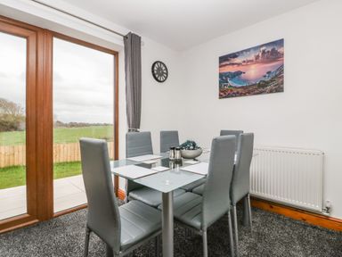 A dining area with six gray chairs around a glass table near large wooden framed windows with a view of a grassy field at Delphine Lodge Meadow View Lodges in Brean