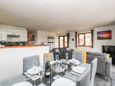 An open plan dining and living area with a glass dining table set for six and a kitchen area in the background at Delphine Lodge Meadow View Lodges in Brean