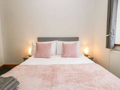A bedroom with a bed with pink pillows and blanket two bedside tables with lamps and a window with grey curtains at Delphine Lodge Meadow View Lodges in Brean