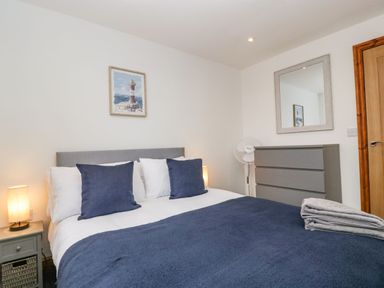 A bedroom with a bed with blue pillows and blanket a nightstand with a lamp a dresser a mirror a standing fan and a painting of a lighthouse on the wall at Delphine Lodge Meadow View Lodges Brean