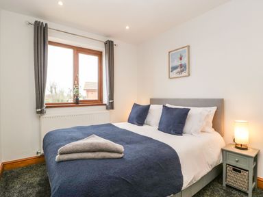 A bedroom with a double bed blue bedding two pillows a bedside table with a lamp and a window with curtains at Delphine Lodge Meadow View Lodges in Brean