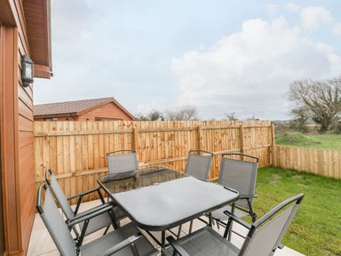 An outdoor patio area with a glass table and six chairs next to a wooden fence with grass beyond at Delphine Lodge Meadow View Lodges in Brean