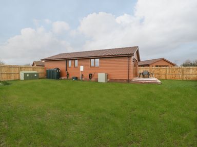 A fenced backyard with green grass and a wooden house with a tiled roof and outdoor seating at Delphine Lodge Meadow View Lodges in Brean