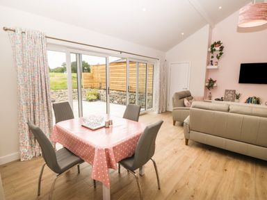 A living room with a dining table covered with a pink polka dot tablecloth and four chairs next to sliding glass doors with patterned curtains at Y Beudy Dinas near Bontnewydd