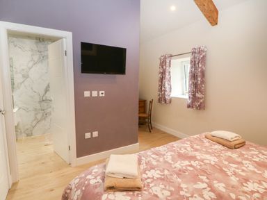 A bedroom with a bed covered in floral bedding and towels on top a wall-mounted tv an open door showing a bathroom and a window with floral curtains at Y Beudy in Dinas near Bontnewydd