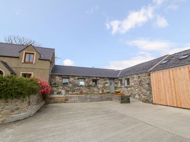 An exterior view of a stone and wooden house with flower beds and a concrete driveway at Y Beudy Dinas near Bontnewydd