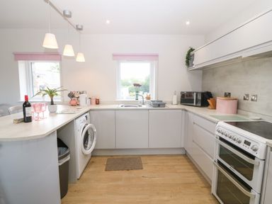A kitchen with white cabinets a washing machine an oven and two windows with pink blinds at Y Beudy in Dinas near Bontnewydd