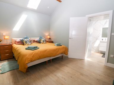 A bedroom with a double bed covered with an orange duvet and pillows next to wooden bedside tables and an open door leading to a bathroom at Y Beudy in Dinas near Bontnewydd