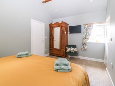 A bedroom with a bed, wooden wardrobe with mirror, green chair with cushions, wall-mounted TV and window at Y Beudy in Dinas near Bontnewydd