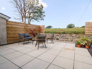 An outdoor patio with a wooden fence stone wall and seating furniture with cushions at Y Beudy in Dinas near Bontnewydd