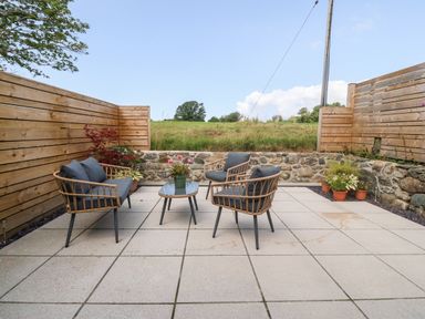 An outdoor patio with rattan furniture and potted plants surrounded by wooden and stone fencing at Y Beudy in Dinas near Bontnewydd