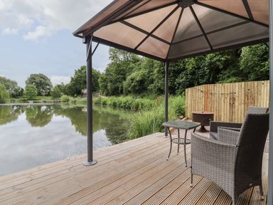 A wooden deck with two wicker chairs a small table and a fire pit overlooking a pond with trees at Maple Lodge at Bridge Lake Farm & Fishery in Chacombe