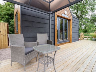An outdoor wooden deck with two wicker chairs and a glass-topped table at Maple Lodge at Bridge Lake Farm & Fishery in Chacombe