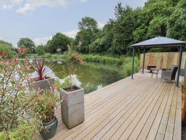 A wooden deck with potted plants a gazebo chairs and a small table next to a pond with trees at Maple Lodge at Bridge Lake Farm & Fishery in Chacombe