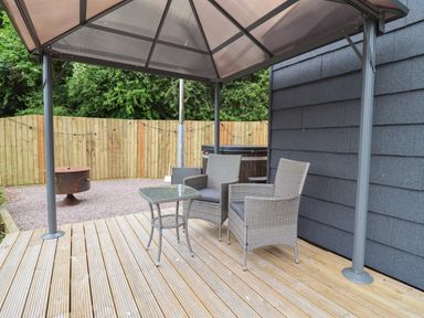 A wooden deck with two wicker chairs and a glass table under a covered structure at Maple Lodge at Bridge Lake Farm Fishery in Chacombe
