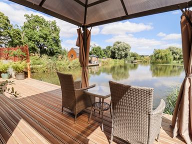 A wooden deck with two wicker chairs and a glass table overlooking a pond with trees and small wooden structures at Maple Lodge at Bridge Lake Farm & Fishery Chacombe