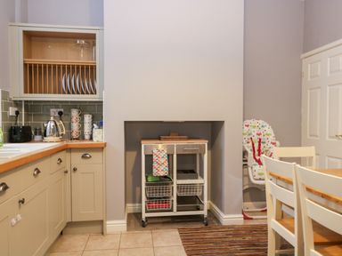 A kitchen with a wooden countertop and cabinets a drying rack with plates a kettle a dining table with chairs and a high chair at 7 Railway Cottages in Newby Bridge