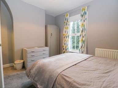 A bedroom with a bed a chest of drawers a wardrobe and a window with floral curtains at 7 Railway Cottages in Newby Bridge