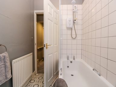 A bathroom with a white bathtub and tiled walls a shower unit a towel on the radiator and an open door at 7 Railway Cottages in Newby Bridge