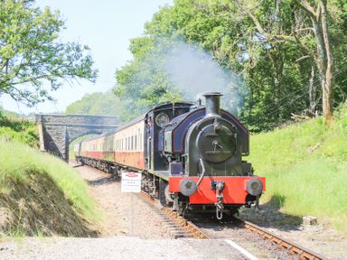 A steam locomotive pulling vintage carriages on a railway track with a stone bridge and green trees at 7 Railway Cottages in Newby Bridge