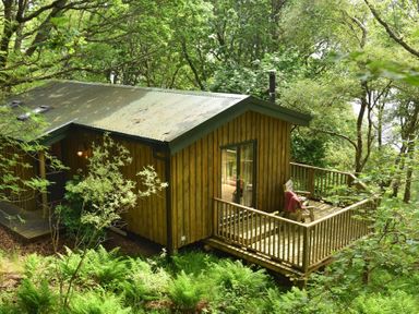 A wooden cabin with a deck and chairs surrounded by trees and greenery at Chapel House in Torridon
