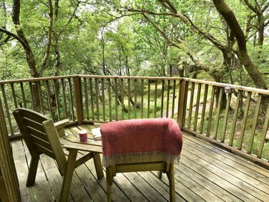 A wooden deck with two chairs a small table a red blanket a book and a mug surrounded by trees at Chapel House in Torridon