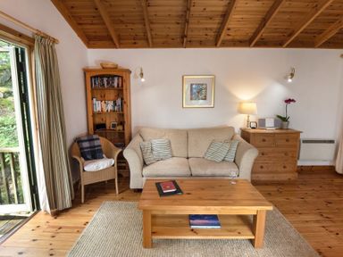 A living room with a beige sofa, a wicker chair, a wooden coffee table, a wooden bookshelf, a wooden chest of drawers, and striped curtains at Chapel House in Torridon