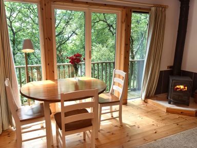 A dining area with a wooden table, white chairs, a vase with red flowers, a wood stove, and large windows showing trees at Chapel House in Torridon