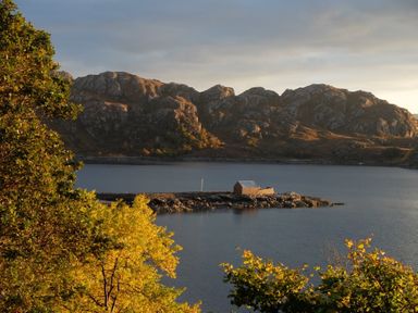 A small stone building on a rocky peninsula surrounded by water with hills and trees in the background at Chapel House in Torridon
