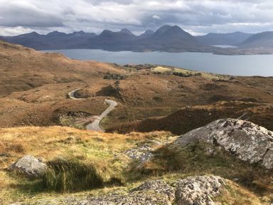 A winding road in a hilly landscape with a lake and mountains in the background at Chapel House in Torridon