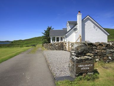 A white house with a chimney next to a stone wall and a gravel driveway at Ardlarach Lodge Cullipool Isle Of Luing