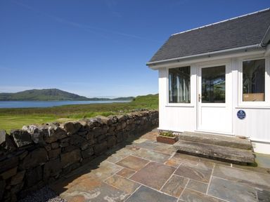Stone patio with steps leading to white door and windows next to stone wall with grassy landscape and water view at Ardlarach Lodge in Cullipool Isle Of Luing
