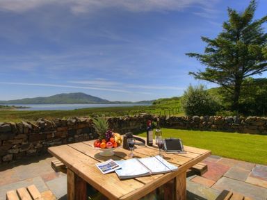 A wooden picnic table with fruit, wine, and maps on a patio overlooking a grassy area and stone wall with water and hills in the background at Ardlarach Lodge in Cullipool Isle Of Luing