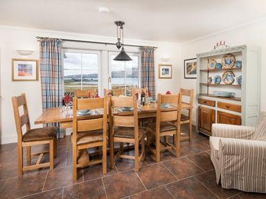 A dining room with a wooden table and chairs a cabinet with plates and a striped armchair at Ardlarach Lodge in Cullipool Isle Of Luing