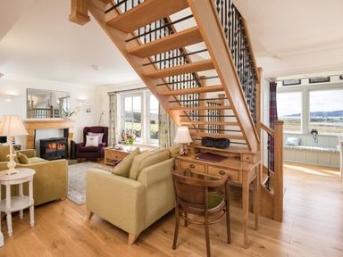A living room with wooden staircase a fireplace armchairs and large windows with a view at Ardlarach Lodge in Cullipool Isle Of Luing