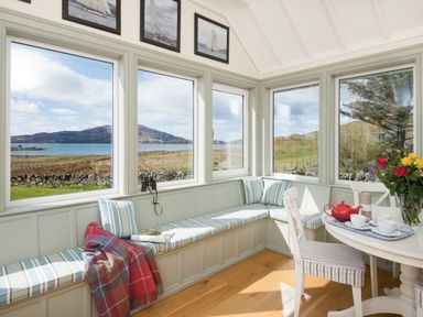 A corner of a room with large windows showing an outdoor landscape a built in bench with cushions a red blanket a table with chairs and a tea set at Ardlarach Lodge Cullipool Isle Of Luing