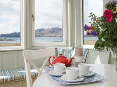 A dining area with a red teapot and three white cups on a tray a striped bench below windows showing a mountain and water view a vase with flowers on the table at Ardlarach Lodge Cullipool Isle Of Luing