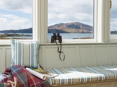 A window seat with striped cushions a plaid blanket a magazine and binoculars with a view of water and hills at Ardlarach Lodge in Cullipool Isle Of Luing