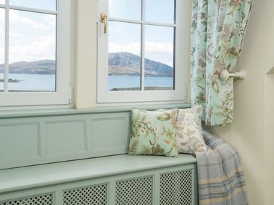 A window seat with floral pillows and a plaid blanket by windows overlooking water and hills at Ardlarach Lodge in Cullipool Isle Of Luing