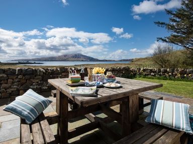 An outdoor wooden table with snacks cups and cushions on benches overlooking a stone wall and a body of water with hills in the background at Ardlarach Lodge in Cullipool Isle Of Luing