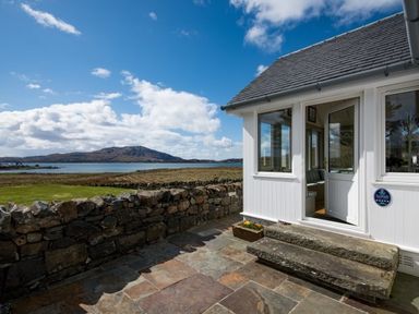An outdoor stone patio with steps leading to a white building and a view of a stone wall grass lake and hills at Ardlarach Lodge Cullipool Isle Of Luing