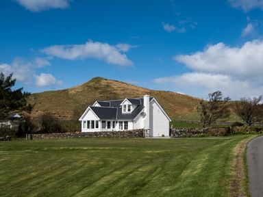 A white house with dark roof near a grassy hill and road at Ardlarach Lodge Cullipool Isle Of Luing